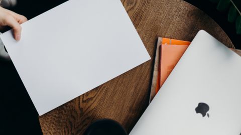 Foto referencial de una mano sosteniendo una hoja de papel en blanco junto a una laptop