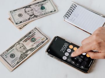 Foto de la mano de una persona usando una calculadora junto a unos billetes y una libreta de apuntes