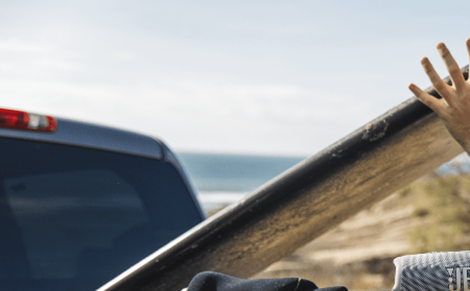 Dos personas cargando una tabla de surf en la parte trasera de una camioneta junto al mar, con un parlante JBL sobre la compuerta.