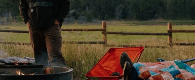 Persona descansando en una silla de camping junto a una fogata, cubierta con una manta de colores.