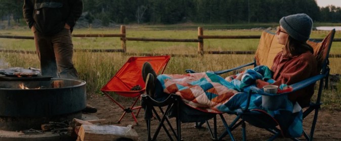 Persona descansando en una silla de camping junto a una fogata, cubierta con una manta de colores.