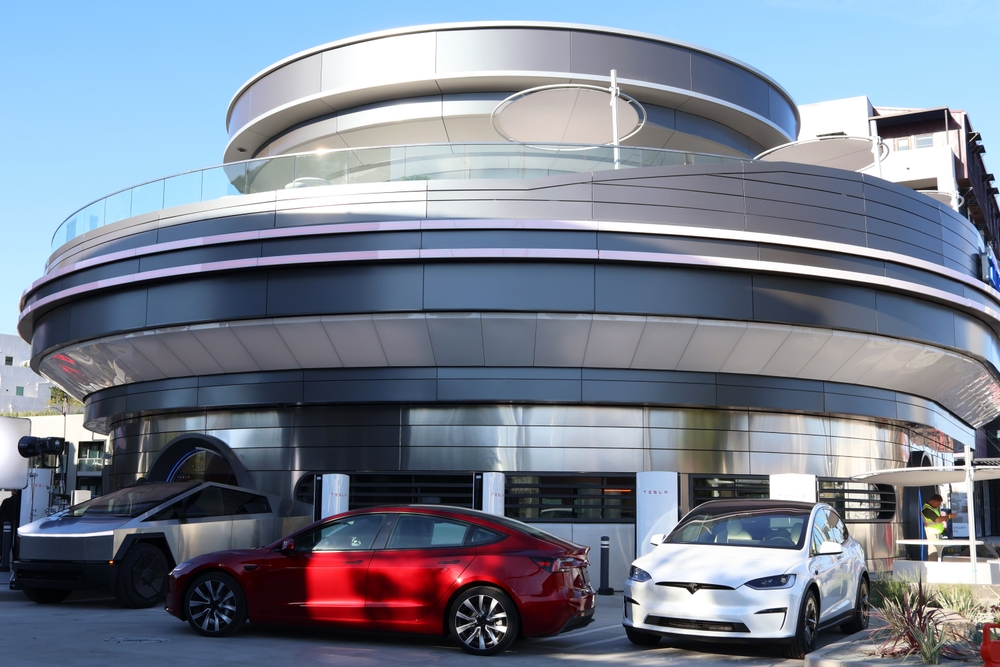 Vista exterior de una tienda Tesla con varios vehículos Tesla, incluyendo un Model 3 rojo, un Model X blanco y un Cybertruck, estacionados frente a la entrada.