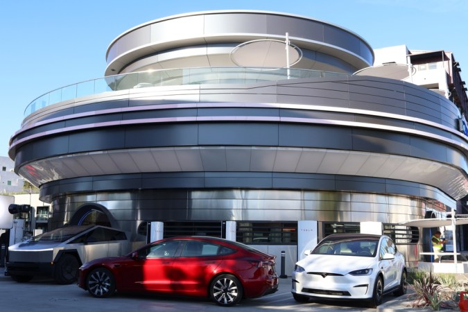 Vista exterior de una tienda Tesla con varios vehículos Tesla, incluyendo un Model 3 rojo, un Model X blanco y un Cybertruck, estacionados frente a la entrada.