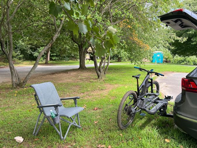 Silla de camping gris colocada en el césped junto a un vehículo con bicicletas montadas en la parte trasera.