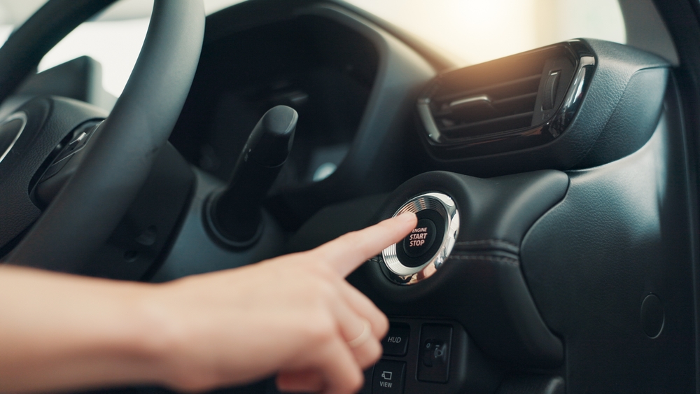 Mano presionando el botón de encendido Start-Stop en el interior de un auto moderno, con el volante y la consola visibles.