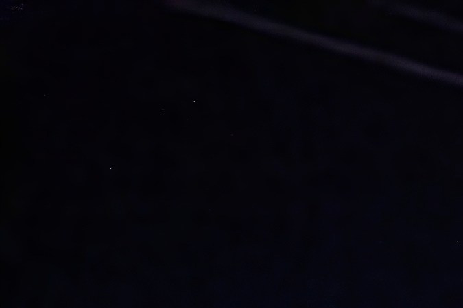 El automóvil autónomo Waymo cruza una intersección por la noche, iluminado por farolas, con peatones caminando y semáforos en el fondo.