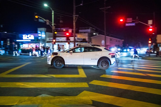 El automóvil autónomo Waymo cruza una intersección por la noche, iluminado por farolas, con peatones caminando y semáforos en el fondo.
