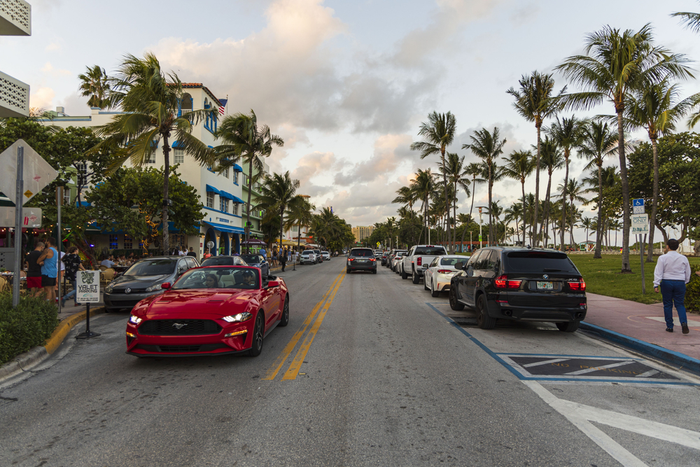 Hermosa vista del paisaje de la calle de Miami Beach. Edificios blancos, coches y palmeras a ambos lados de la carretera asfaltada sobre el cielo azul y el fondo de nubes blancas