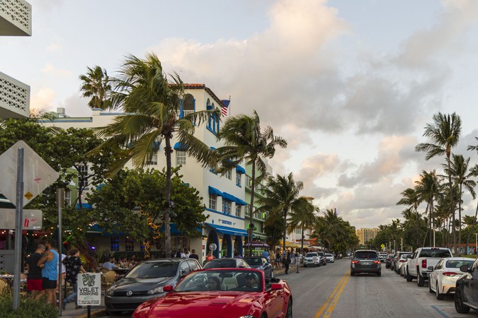 Hermosa vista del paisaje de la calle de Miami Beach. Edificios blancos, coches y palmeras a ambos lados de la carretera asfaltada sobre el cielo azul y el fondo de nubes blancas