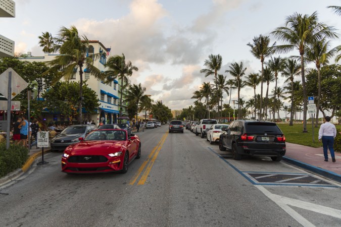 Hermosa vista del paisaje de la calle de Miami Beach. Edificios blancos, coches y palmeras a ambos lados de la carretera asfaltada sobre el cielo azul y el fondo de nubes blancas