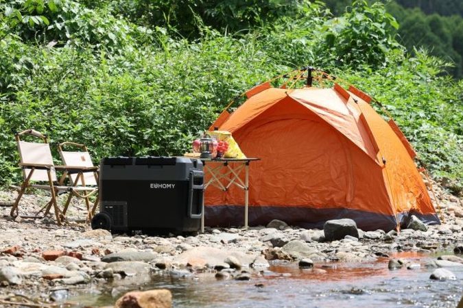 Campamento junto a un arroyo con una tienda de campaña naranja, sillas plegables y una nevera portátil Euhomy.