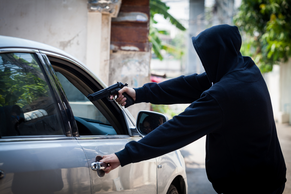 Ladron con capucha apunta con pistola a conductor dentro de auto gris en calle.