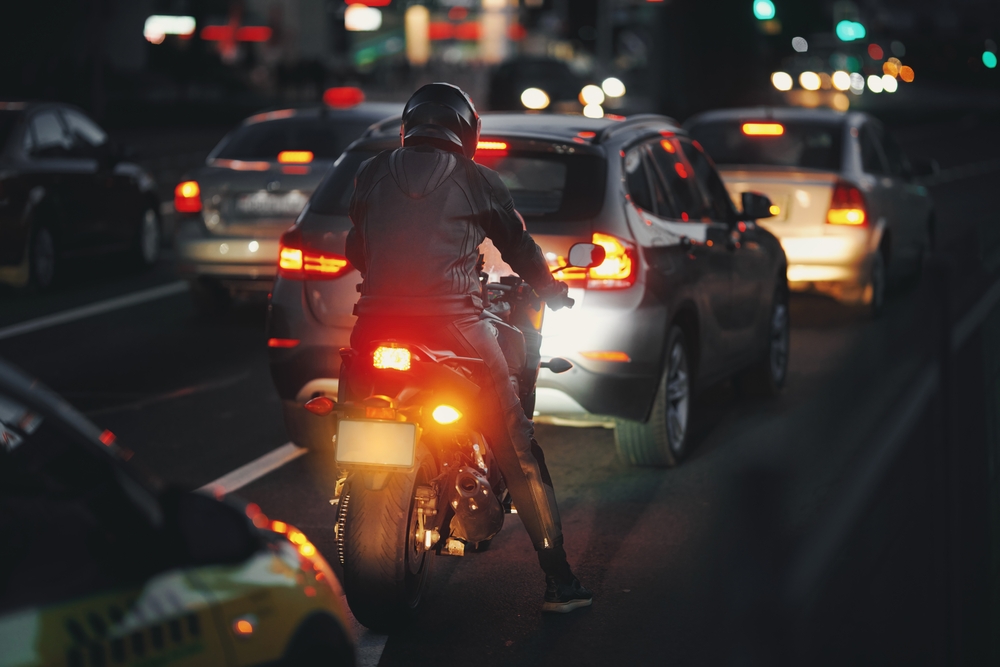 Motociclista en tráfico nocturno ahorrando gasolina con velocidad eficiente entre autos en ciudad.