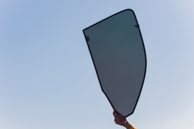 Persona sosteniendo un parasol negro de malla para ventana de carro, con el cielo azul de fondo.