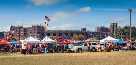 Una escena animada de tailgating con carpas coloridas, banderas y personas reunidas alrededor de vehículos en un campo de hierba, con un edificio de estadio de fondo bajo un cielo azul despejado.
