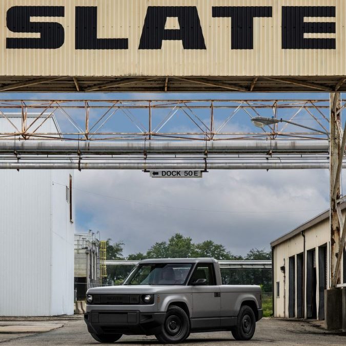 Camioneta eléctrica Slate gris estacionada frente a una estructura industrial con el letrero grande de ‘SLATE’.