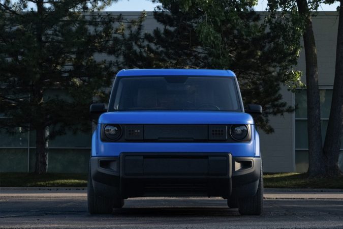 Vista frontal de una camioneta eléctrica Slate azul estacionada al aire libre frente a árboles y un edificio
