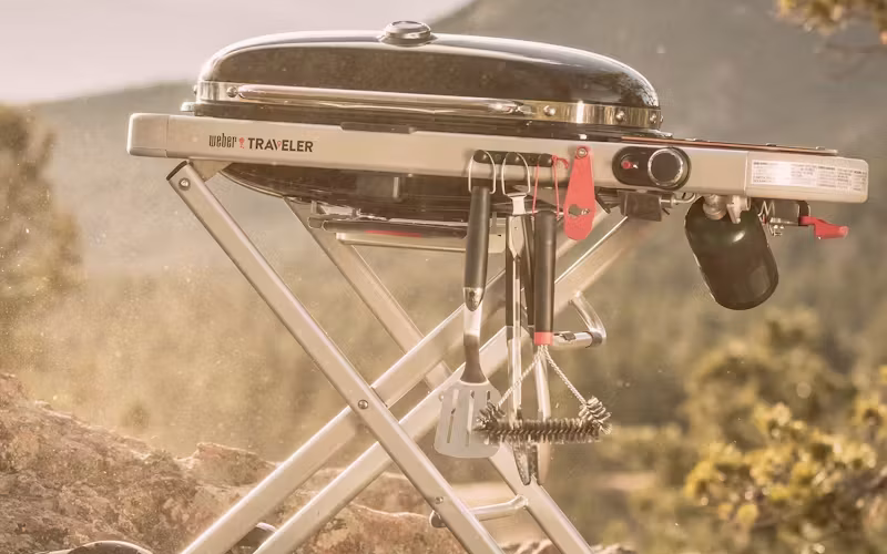 Persona cocinando al aire libre en una parrilla portátil Weber Traveler junto a un vehículo.