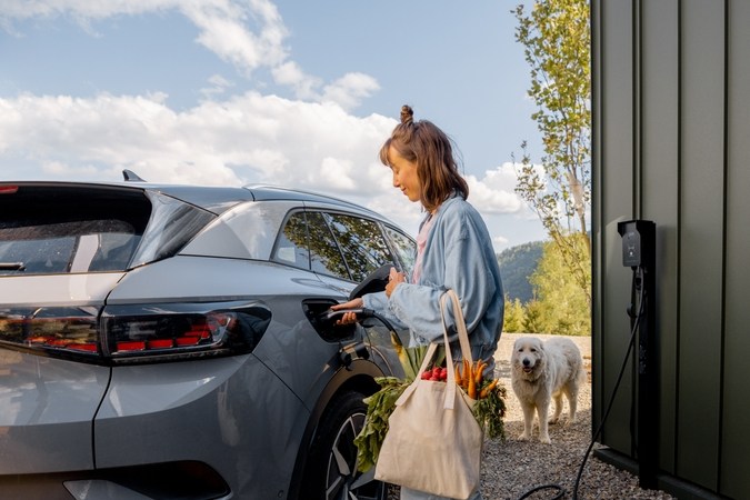 Mujer joven enchufando un cargador en el vehículo eléctrico cerca de su casa al aire libre. Concepto de tecnologías modernas, coches eléctricos y estilo de vida sostenible