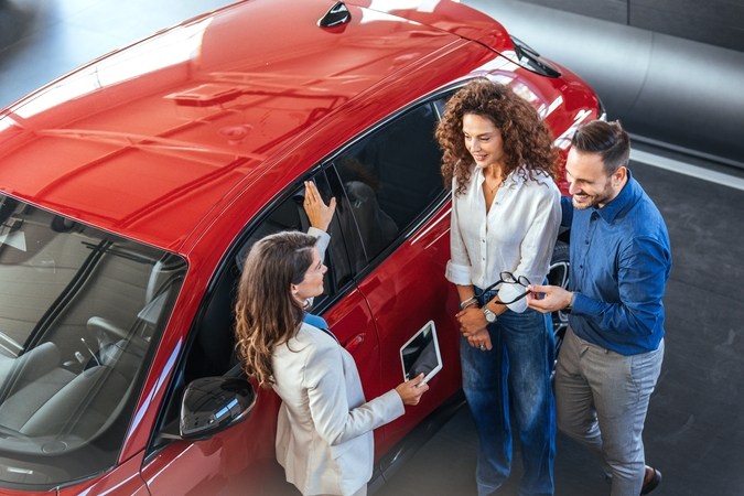 Una pareja joven discute las opciones de automóviles con un Representante del concesionario en una sala de exposición moderna, destacando la emoción y la toma de decisiones en la compra de un nuevo vehículo.