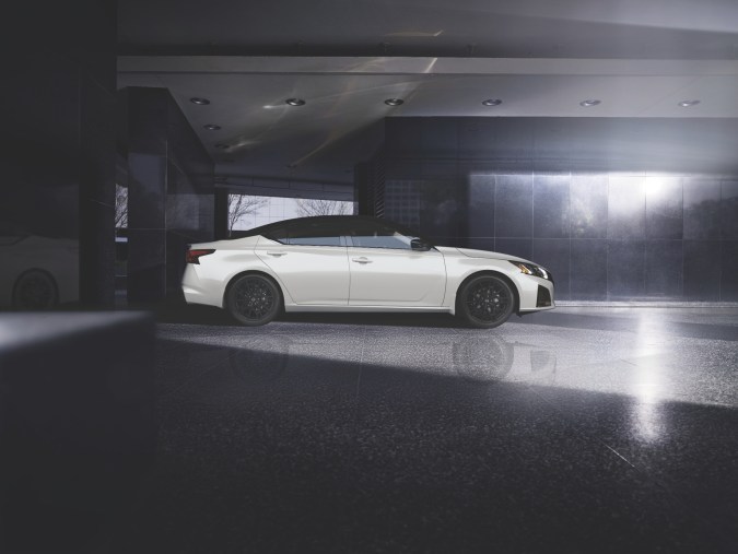 Nissan Altima 2026 blanco de perfil en estacionamiento cubierto, llantas negras y silueta deportiva iluminada por luz lateral.