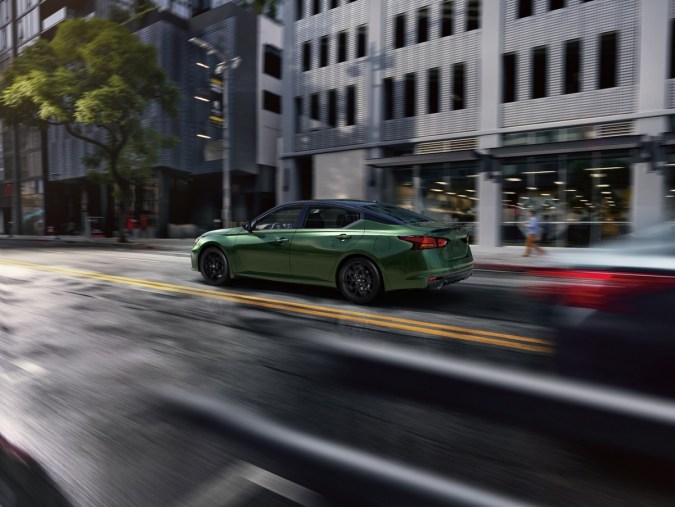 Nissan Altima verde 2026 en movimiento por ciudad, llantas negras, techo oscuro y fondo urbano desenfocado que resalta su diseño dinámico.