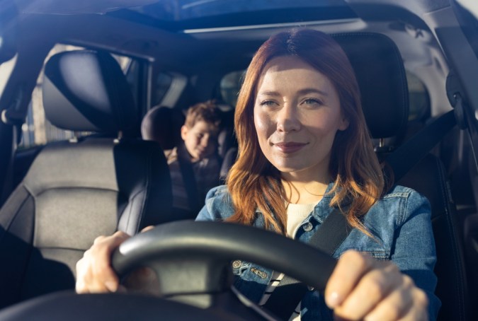 Mujer sonriendo mientras conduce el coche, con el niño sentado cómodamente en el asiento trasero. La luz del sol ilumina su viaje