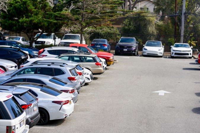 Un ocupado estacionamiento al aire libre lleno de una variedad de vehículos estacionados, incluidos SUV, sedanes y autos eléctricos, rodeado de árboles en un entorno suburbano - California