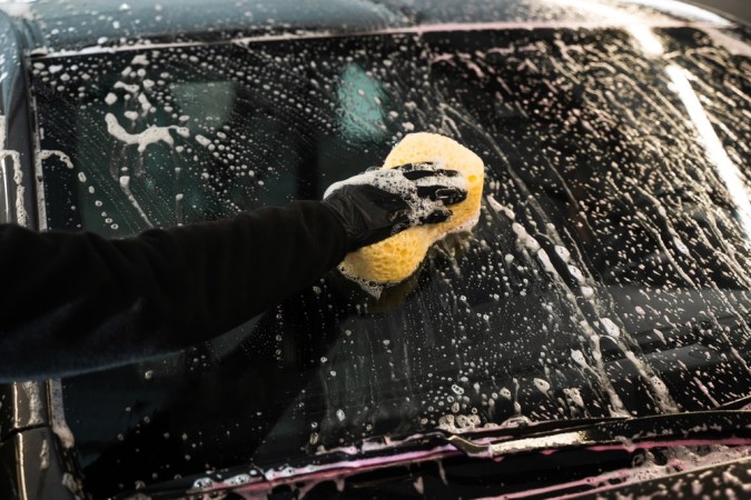 Los espumones caen en cascada por la luna del vehículo. El limpiador de coches está lavando el parabrisas con esponja y detergente de espuma en el servicio de lavado de coches