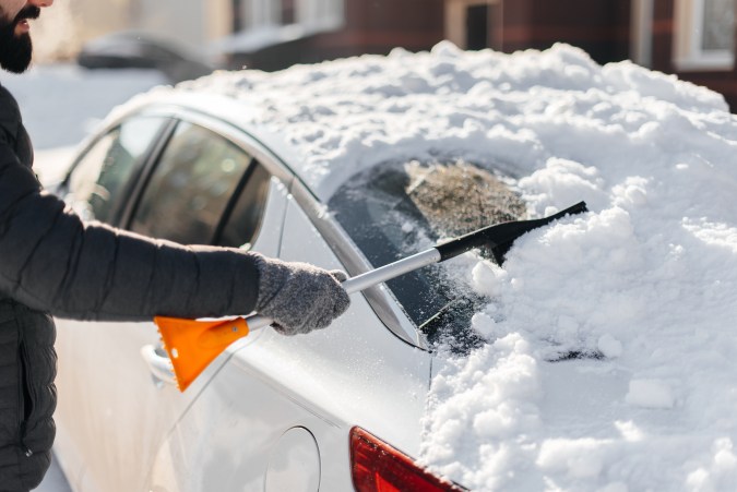 Un joven limpia su auto después de una nevada en un día soleado y helado. Limpiando y limpiando el auto de la nieve en un día de invierno. Nevada y una fuerte tormenta de nieve en invierno.