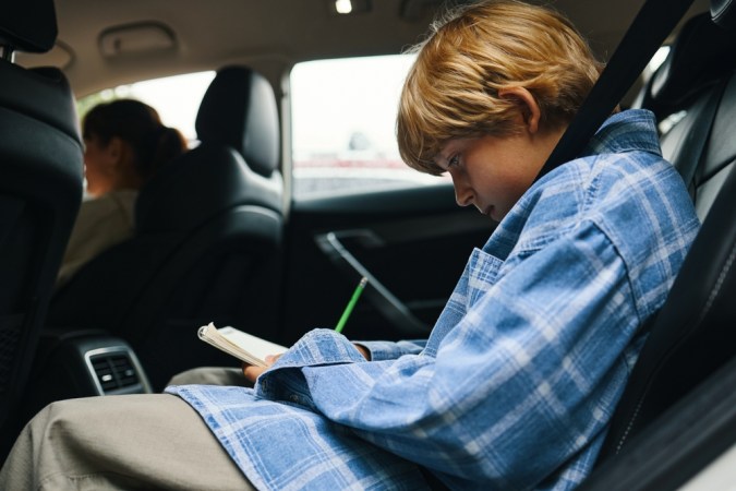 Niño caucásico sentado en el asiento trasero del coche escribiendo en el cuaderno durante el viaje por carretera familiar, mujer de mediana edad visible en la conducción del asiento delantero, ambos con cinturones de seguridad, expresiones enfocadas