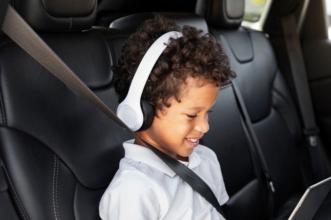 Niño viajando en auto compartiendo una tableta de juegos de mano. Preteen, un chico afroamericano con audífonos inalámbricos usando tableta digital sentado en el asiento trasero del auto