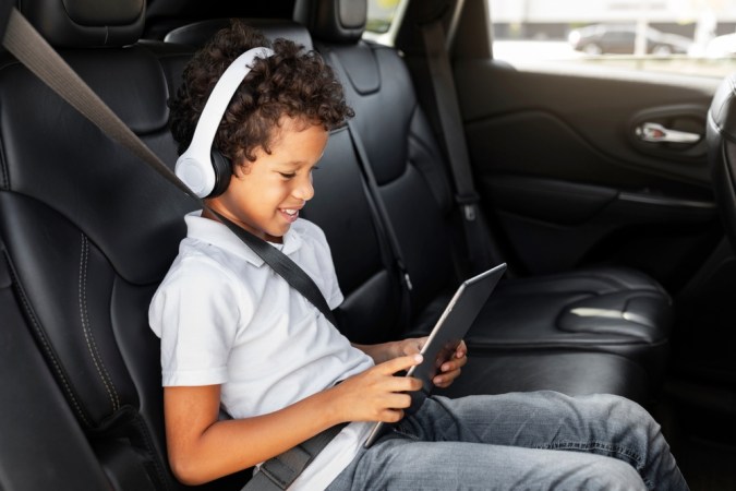 Niño viajando en auto compartiendo una tableta de juegos de mano. Preteen, un chico afroamericano con audífonos inalámbricos usando tableta digital sentado en el asiento trasero del auto