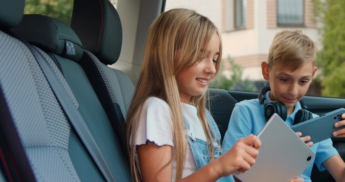 Muchacha atractiva jugando una tablet y un smartphone en el coche. Niños que usan dispositivos para juegos y entretenimiento, viajando en auto.