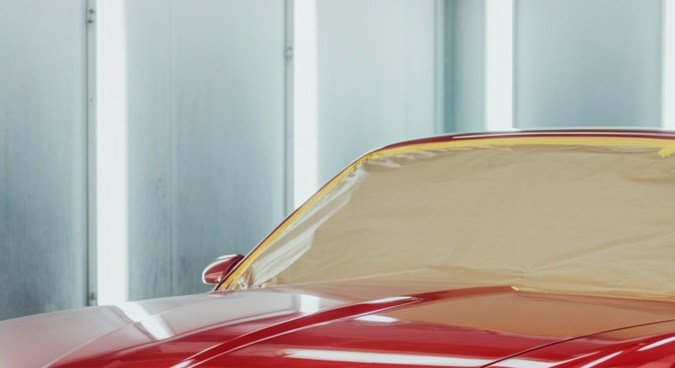 Garaje de un taller de pintura de coches. Cámara lateral gran angular del suelo. Vemos una pared azul real sencilla al fondo. Delante de la pared vemos a un pintor sonriente, con guantes y monos azules, tela de franela amarilla en la mano, el cuerpo inclinado, mirando