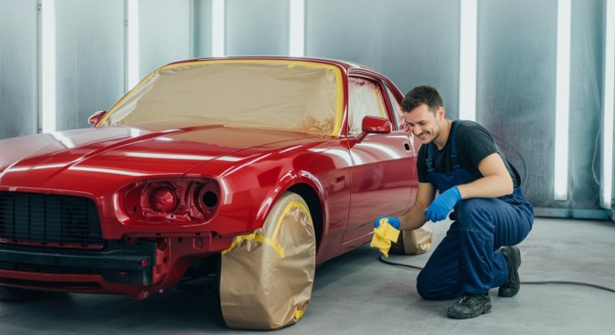 Garaje de un taller de pintura de coches. Cámara lateral gran angular del suelo. Vemos una pared azul real sencilla al fondo. Delante de la pared vemos a un pintor sonriente, con guantes y monos azules, tela de franela amarilla en la mano, el cuerpo inclinado, mirando
