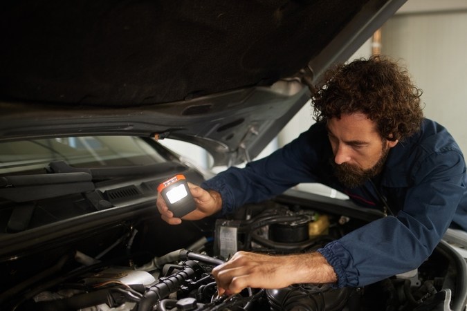 La mecánica se centra en diagnosticar y solucionar problemas del motor bajo el capó de un automóvil, utilizando una luz para inspeccionar piezas en un entorno de taller profesional