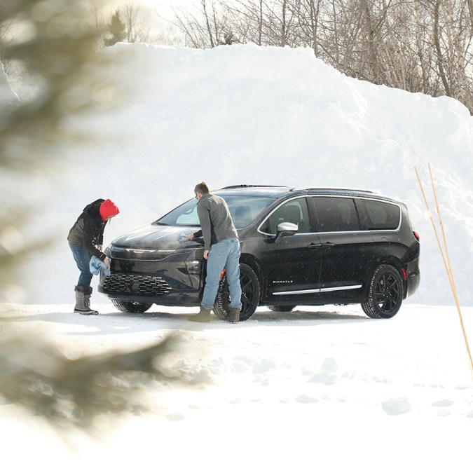 Pareja limpiando nieve del nuevo Chrysler Pacifica 2027 en paisaje invernal, mostrando diseño elegante y robusto.