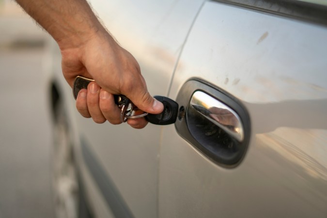 Una persona está abriendo la puerta de un automóvil con una llave, mostrando un primer plano de la mano y la llave mientras está de pie en un estacionamiento durante el día.