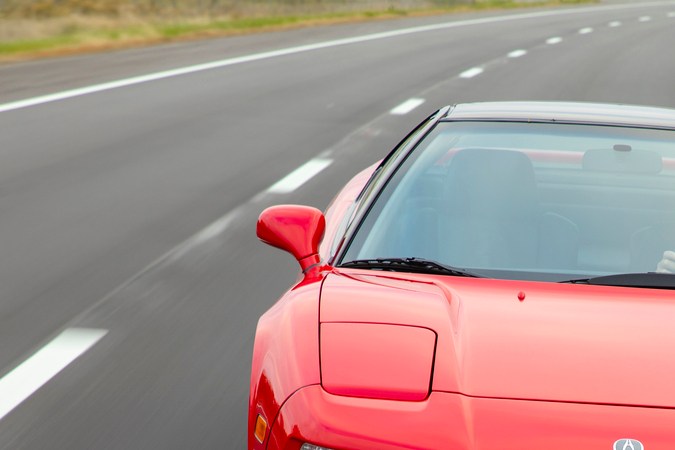 Acura NSX 1991 rojo acelerando en carretera, perfil frontal con motor central y líneas agresivas del superdeportivo japonés.