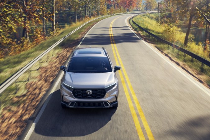 Honda CR-V Sport Touring Hybrid 2026. plateado en carretera otoñal boscosa, SUV compacto familiar de diseño moderno.