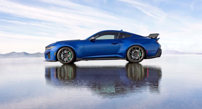 Ford Mustang Dark Horse SC azul de perfil sobre superficie mojada, con alerón trasero y rines deportivos, fotografía promocional para entusiastas de autos deportivos.