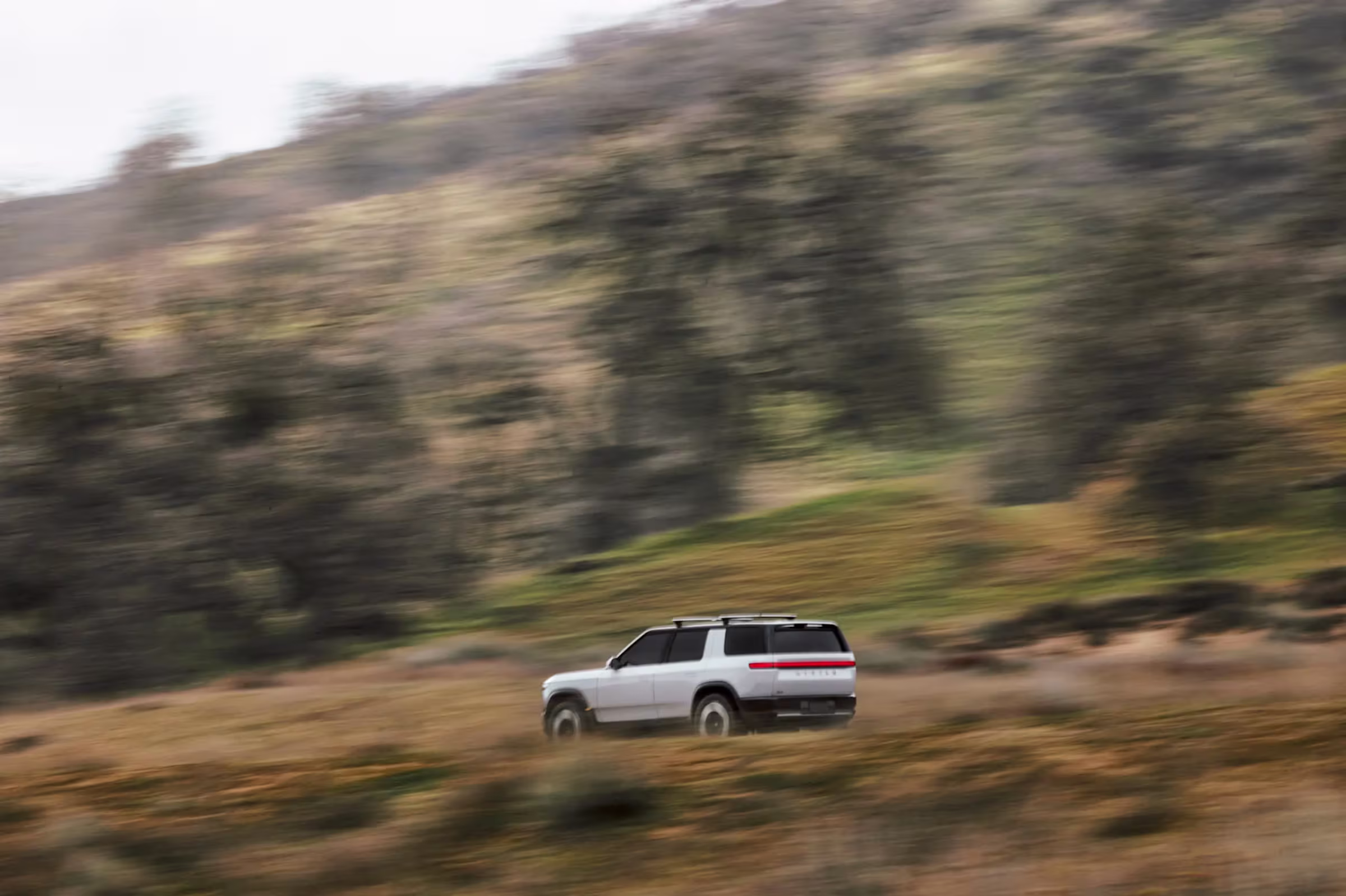 Rivian R2 2027 en perfil lateral sobre colina verde, SUV compacto eléctrico para aventuras y ciudad