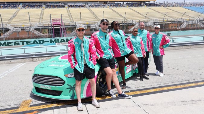 Homestead-Miami Speedway con grupo diverso en uniformes rosados y verde junto a auto Ford Mustang tropical. Día de prensa NASCAR Miami.