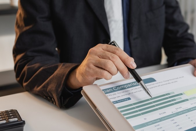 Un hombre de negocios en un traje está calculando los detalles del préstamo, revisando documentos financieros y sosteniendo las llaves del automóvil cerca de una calculadora y modelos de automóviles de juguete, que representan los servicios de préstamos y finanzas para automóviles.