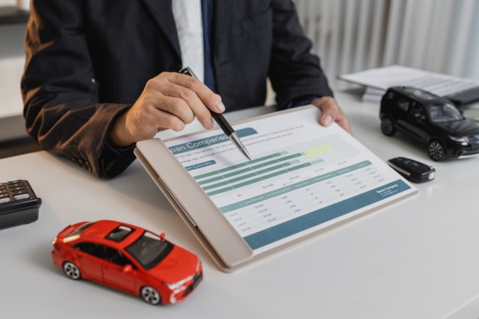 Un hombre de negocios en un traje está calculando los detalles del préstamo, revisando documentos financieros y sosteniendo las llaves del automóvil cerca de una calculadora y modelos de automóviles de juguete, que representan los servicios de préstamos y finanzas para automóviles.