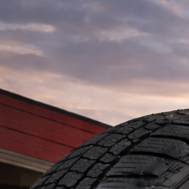Primer plano de neumático Firestone frente a taller automotriz al atardecer, con camioneta y SUV sobre asfalto mojado iluminado por luz cálida.