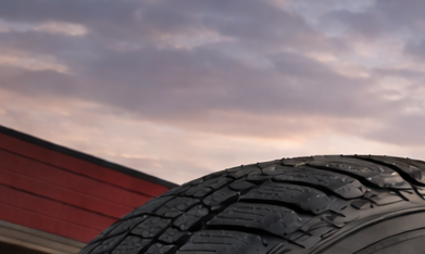 Primer plano de neumático Firestone frente a taller automotriz al atardecer, con camioneta y SUV sobre asfalto mojado iluminado por luz cálida.