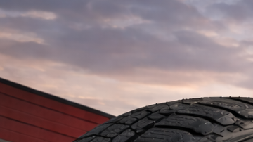 Primer plano de neumático Firestone frente a taller automotriz al atardecer, con camioneta y SUV sobre asfalto mojado iluminado por luz cálida.