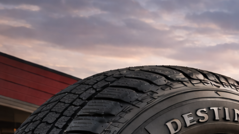 Primer plano de neumático Firestone frente a taller automotriz al atardecer, con camioneta y SUV sobre asfalto mojado iluminado por luz cálida.